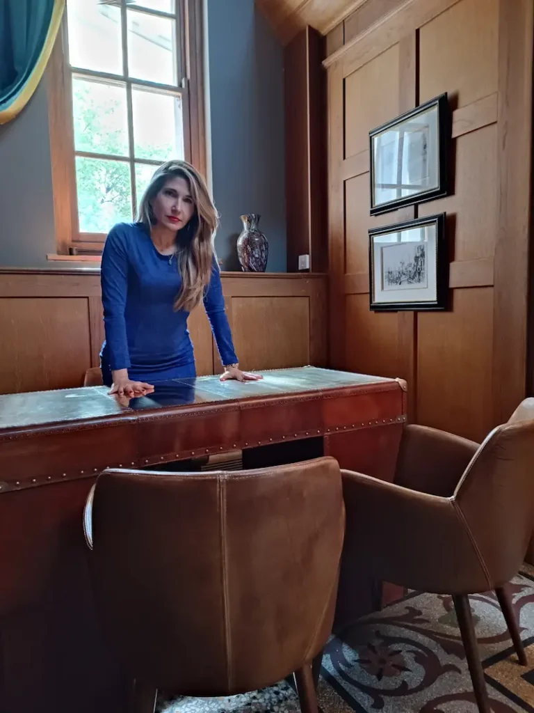 Retrato de Mistress Bibianne, mentora de Dominación Femenina Sagrada, de pie con actitud de absoluta autoridad detrás de un clásico escritorio de cuero en un elegante despacho de madera, vistiendo un vestido azul y mirando firmemente a la cámara.